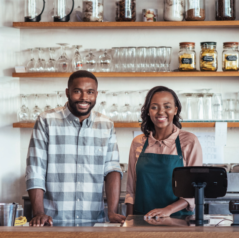 A smiling pair behind a café counter, standing in front of shelves filled with glassware and jars.