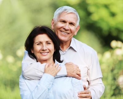 Smiling couple embracing outdoors. Woman in front, man's arms around her. Green background.