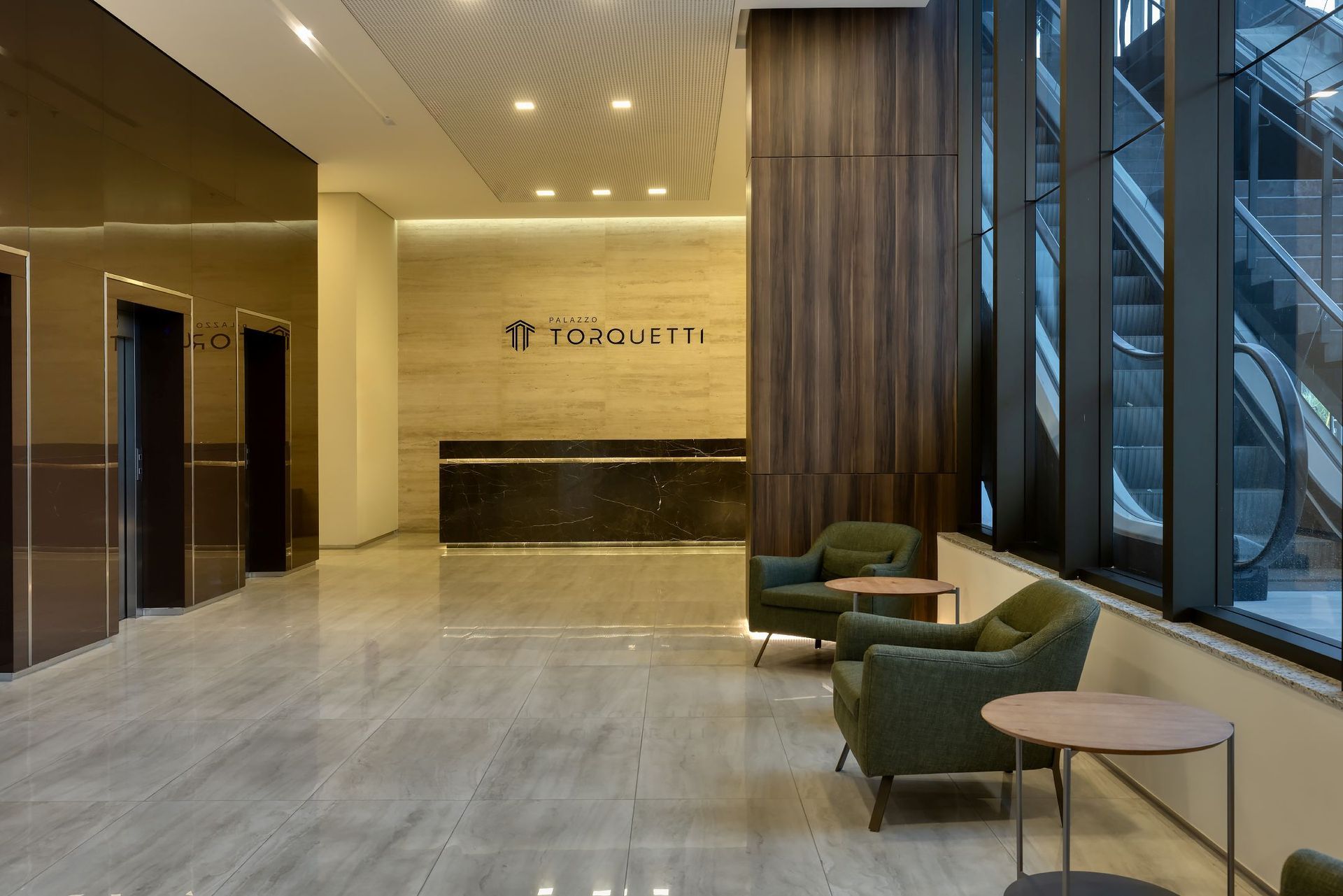 Um lobby com balcão de recepção, cadeiras, mesas e escadas rolantes.