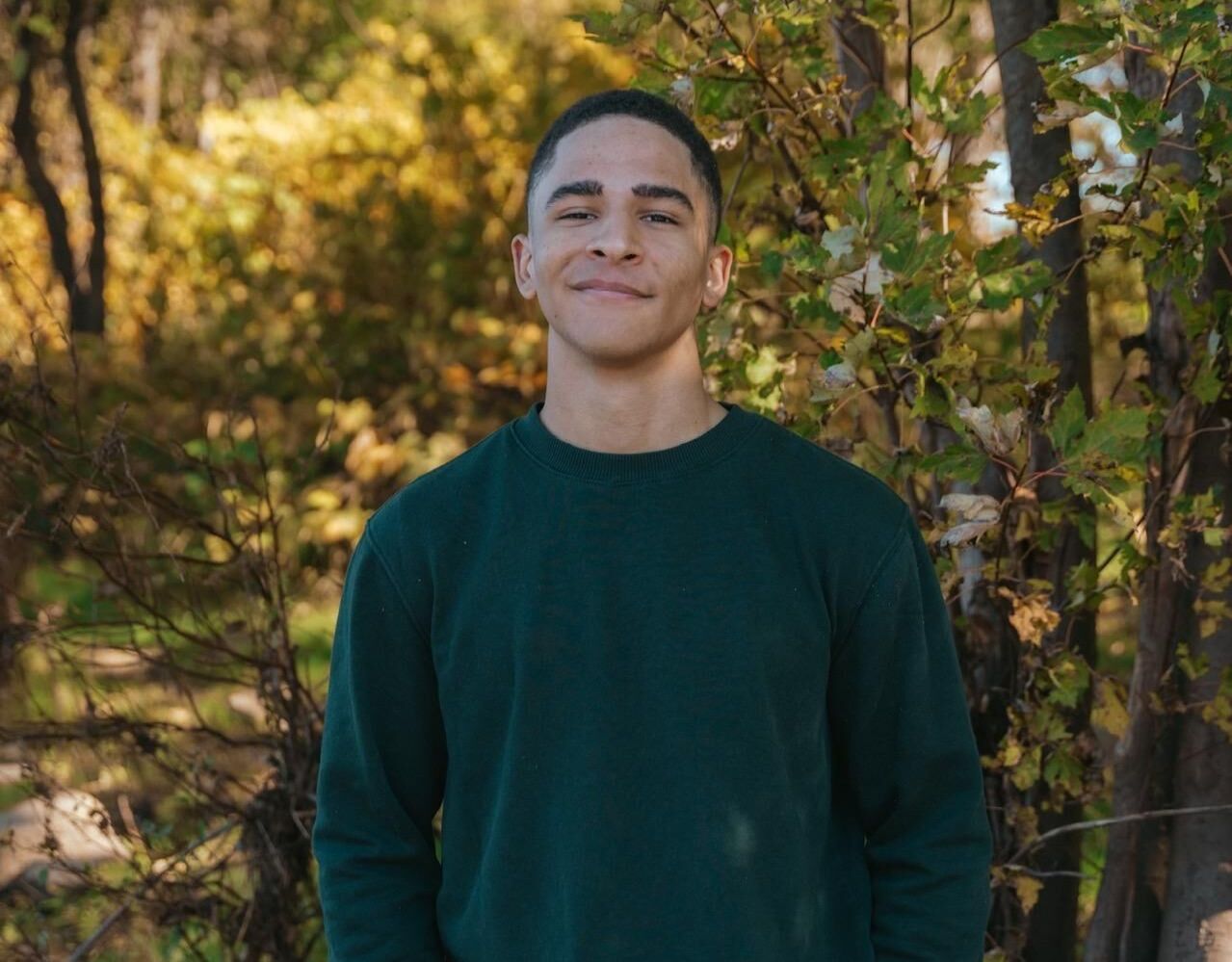 A young man in a green sweater is standing in the woods.