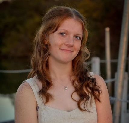 A young woman with long red hair and a necklace is smiling for the camera.