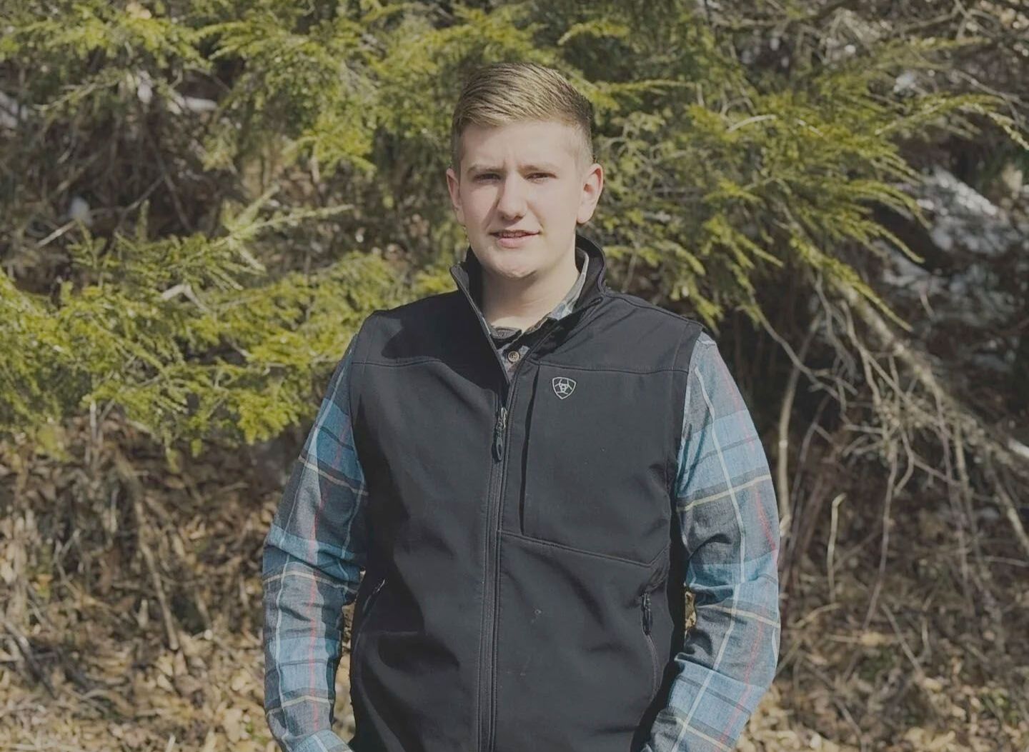 A young man wearing a black vest and a plaid shirt is standing in the woods.