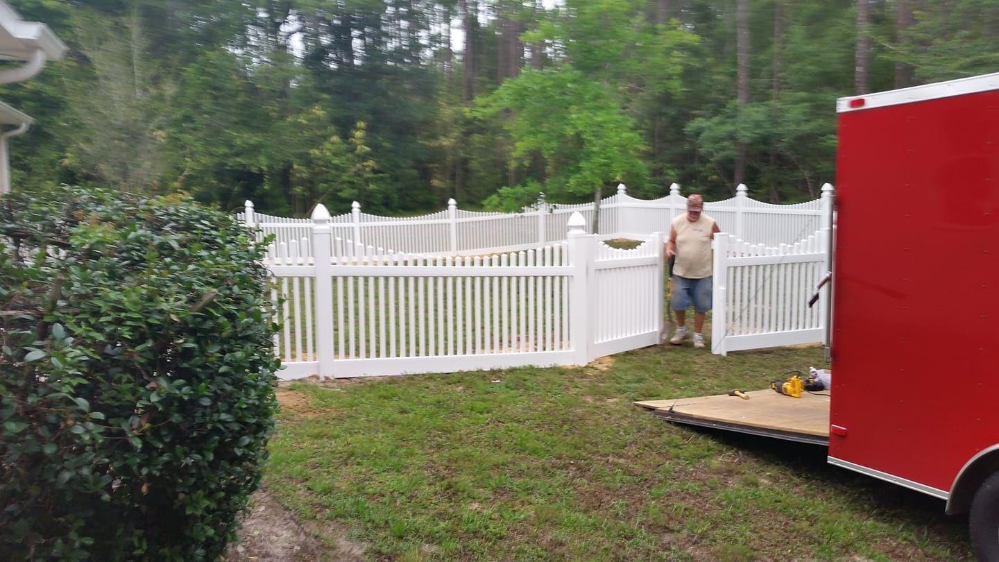 Man stands near a partially built white picket fence in a grassy backyard, red trailer on the right.