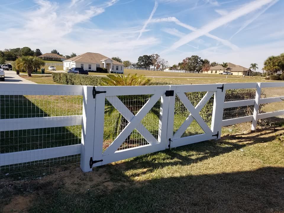 White gate with an X design, part of a white fence, in front of a house on a sunny day.