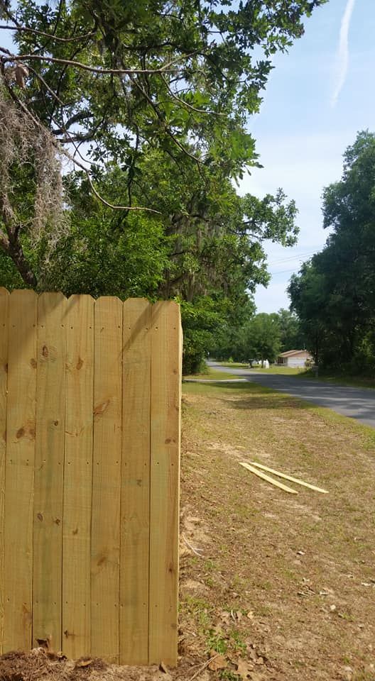 Wooden fence borders a grassy area next to a road, trees in the background, blue sky.