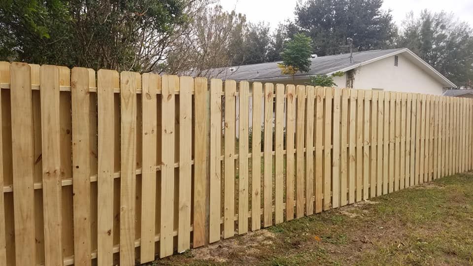 Wooden fence in a yard, light brown, vertical planks, running along a grassy area, house in the background.