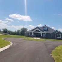 Asphalt driveway leads to a gray house with a blue sky background.