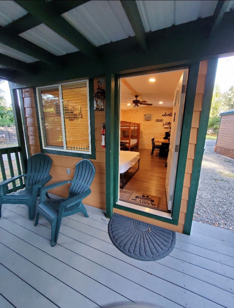 A cabin porch with two green chairs, a door revealing a room with bunk beds, and a gray doormat on light-colored wood floor.