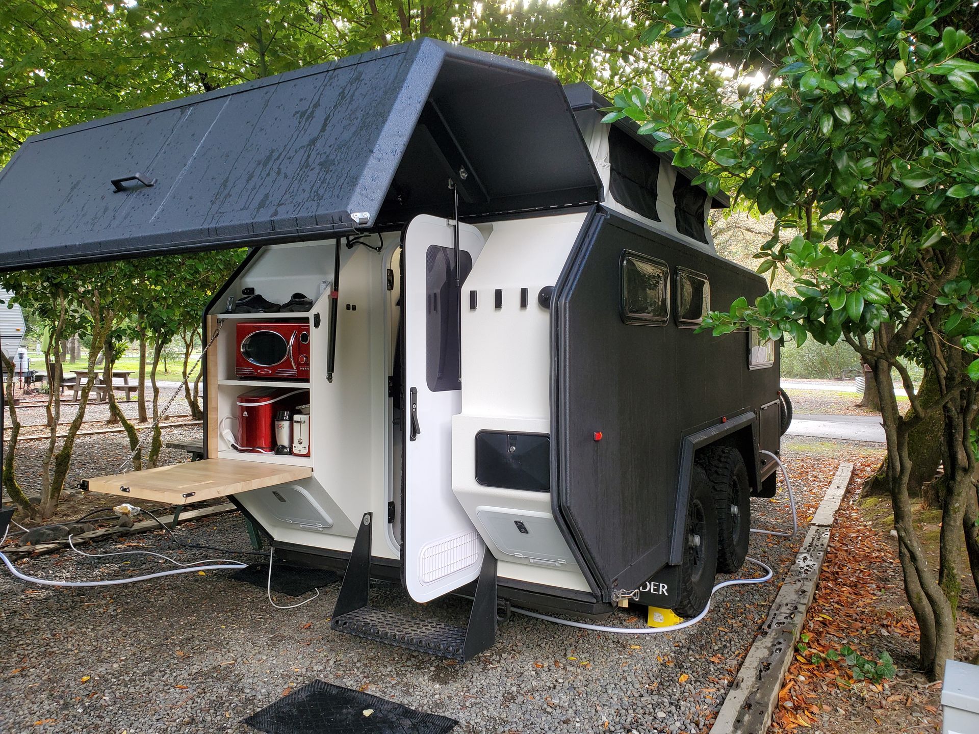 A black and white off-road camper trailer parked in a wooded area with its side hatch open to reveal a kitchen setup.
