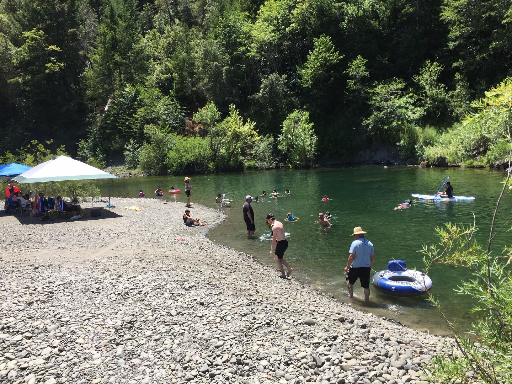 People enjoy a sunny day at a gravel riverbank with trees, a canopy, a paddleboarder, and individuals wading in the water.