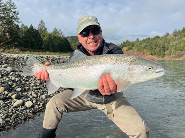 A person wearing a cap and sunglasses holds a large, silvery fish while crouching by a rocky riverbank.