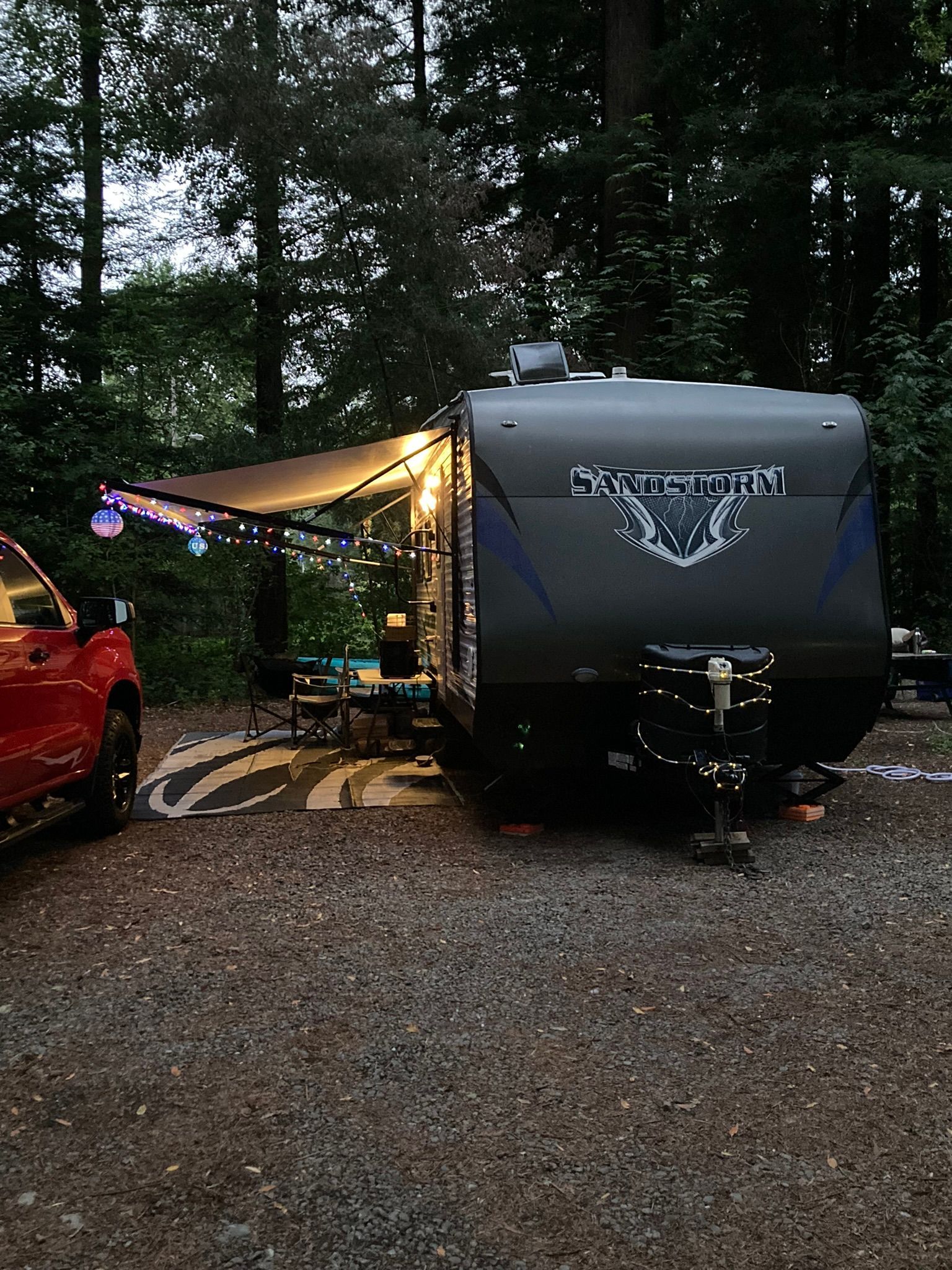 A dark gray Keystone travel trailer parked in a wooded campsite with a lit awning and a red truck nearby.