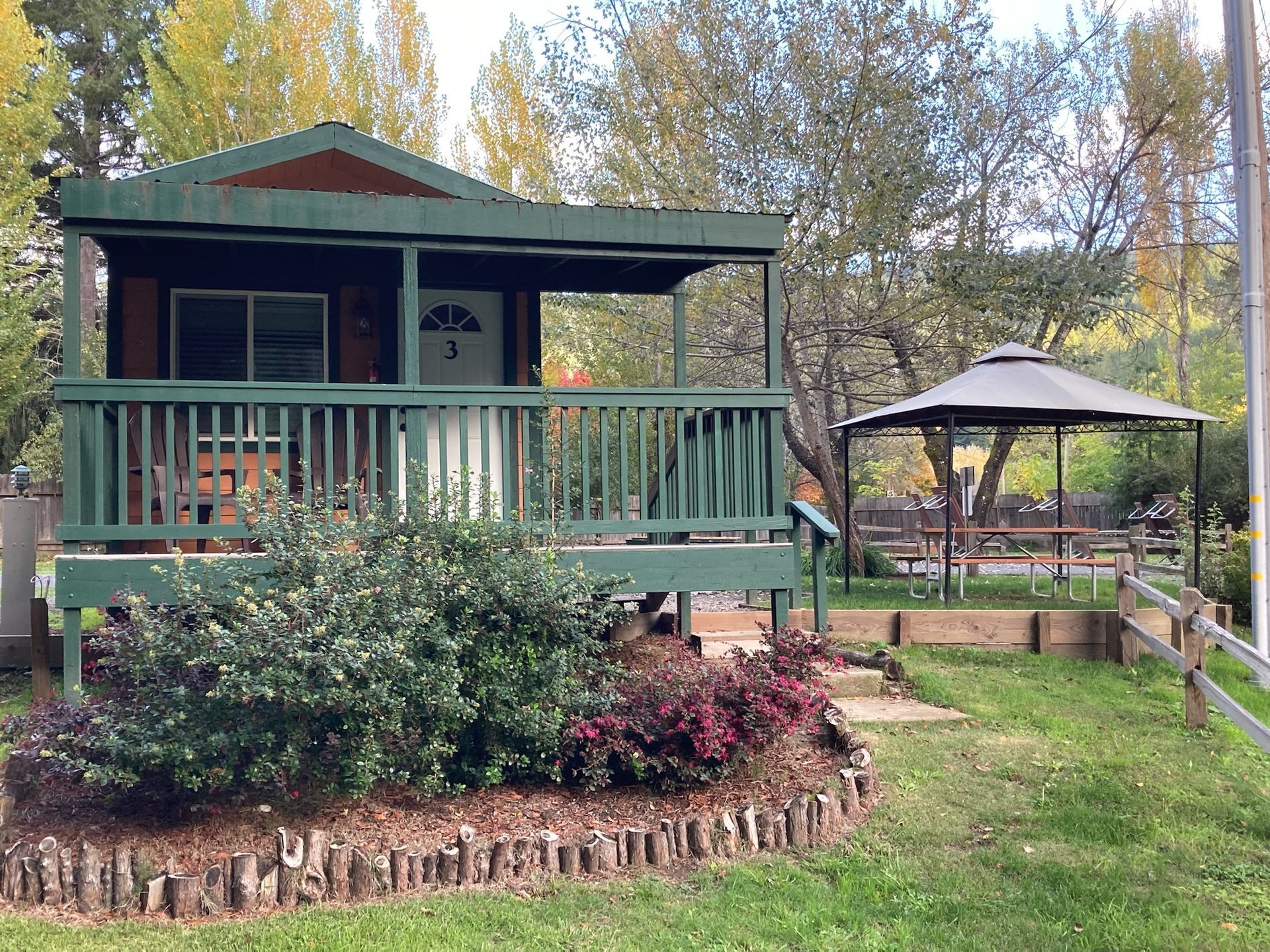A small, green wooden cabin with a front porch sits in a grassy area next to a brown gazebo under autumn trees.