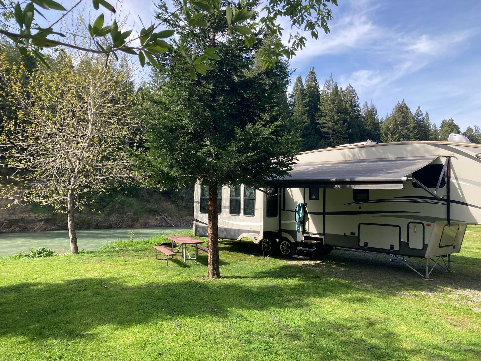 A large beige recreational vehicle parked on a green lawn by a river, shaded by trees under a blue sky.