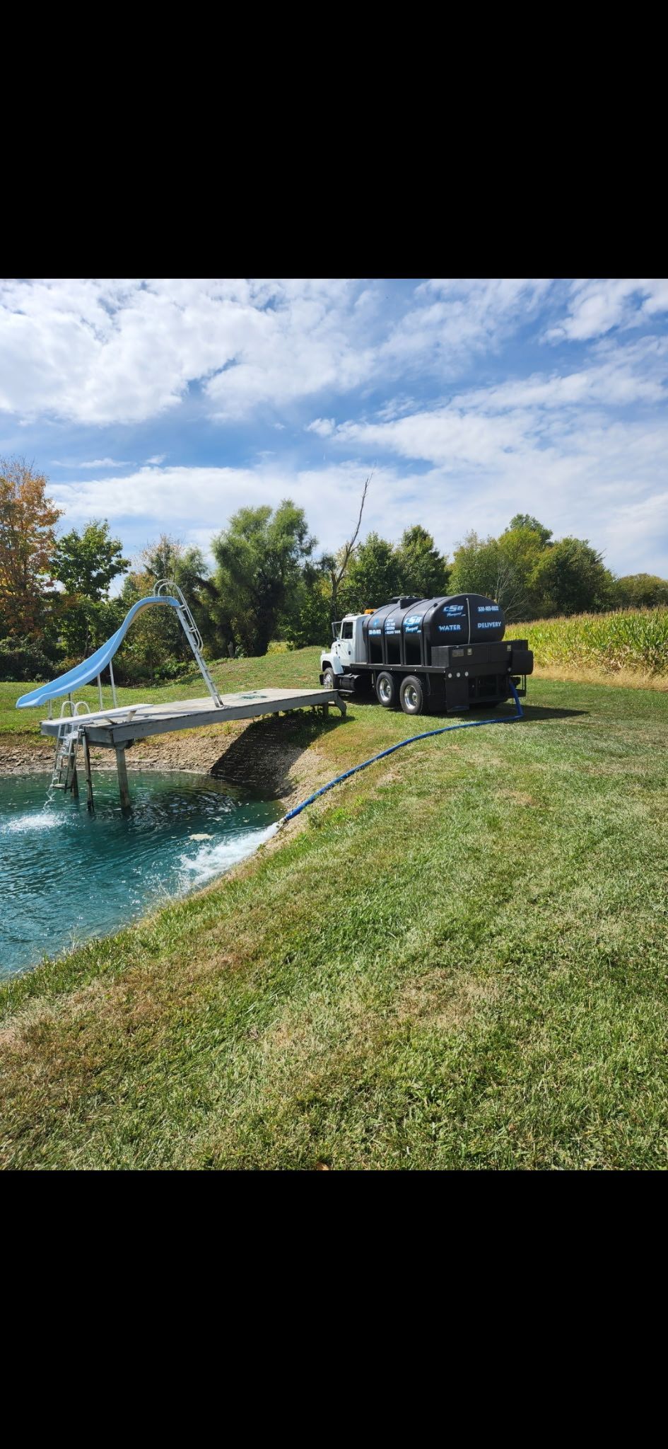 A truck near a pond is discharging water into the pond via a ramp, in a grassy field.