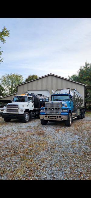 Two tanker trucks parked in front of a barn. One truck is white, the other is blue. The ground is gravel.