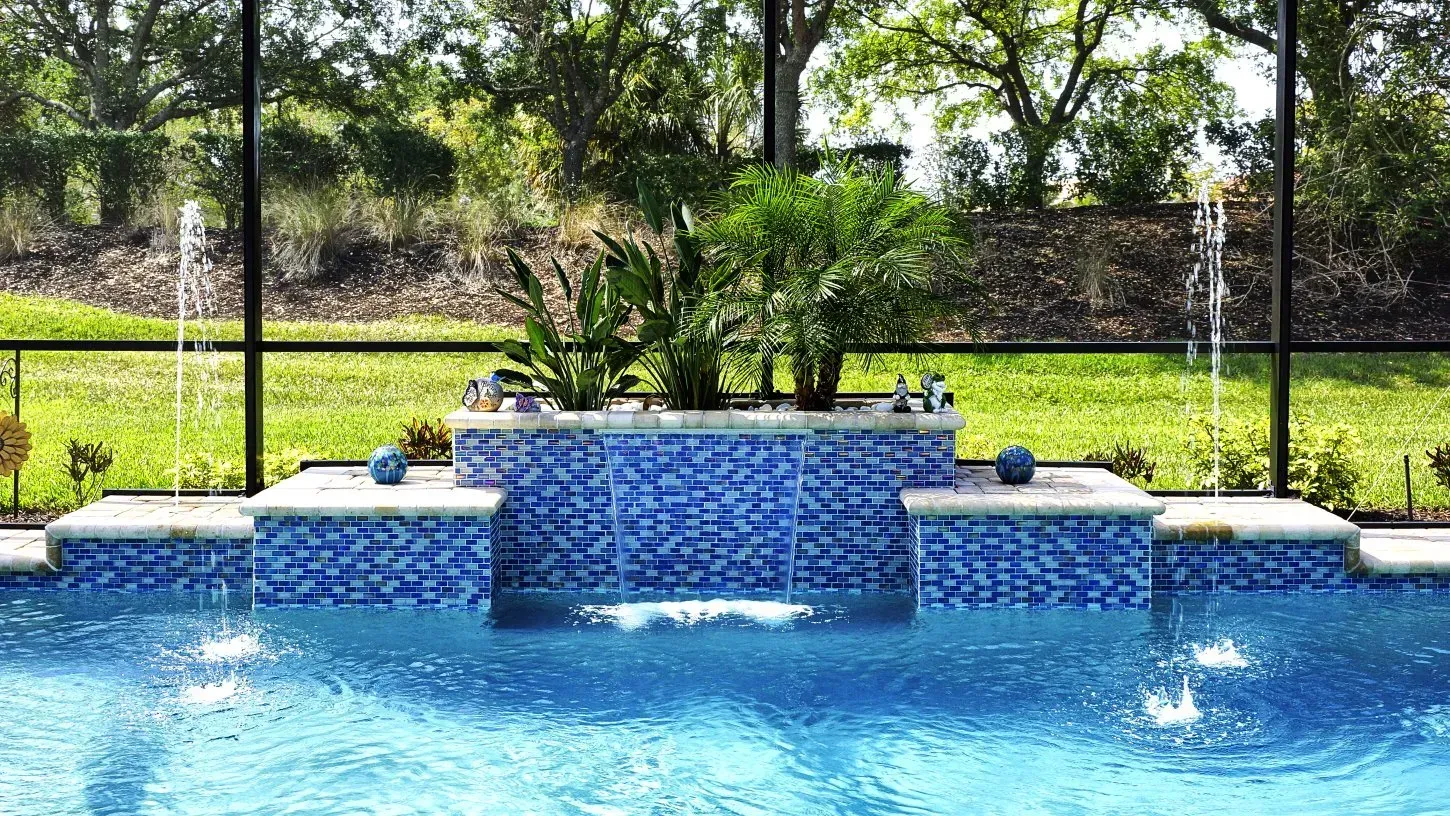 Blue tiled pool with cascading waterfall and fountains, lush greenery backdrop.