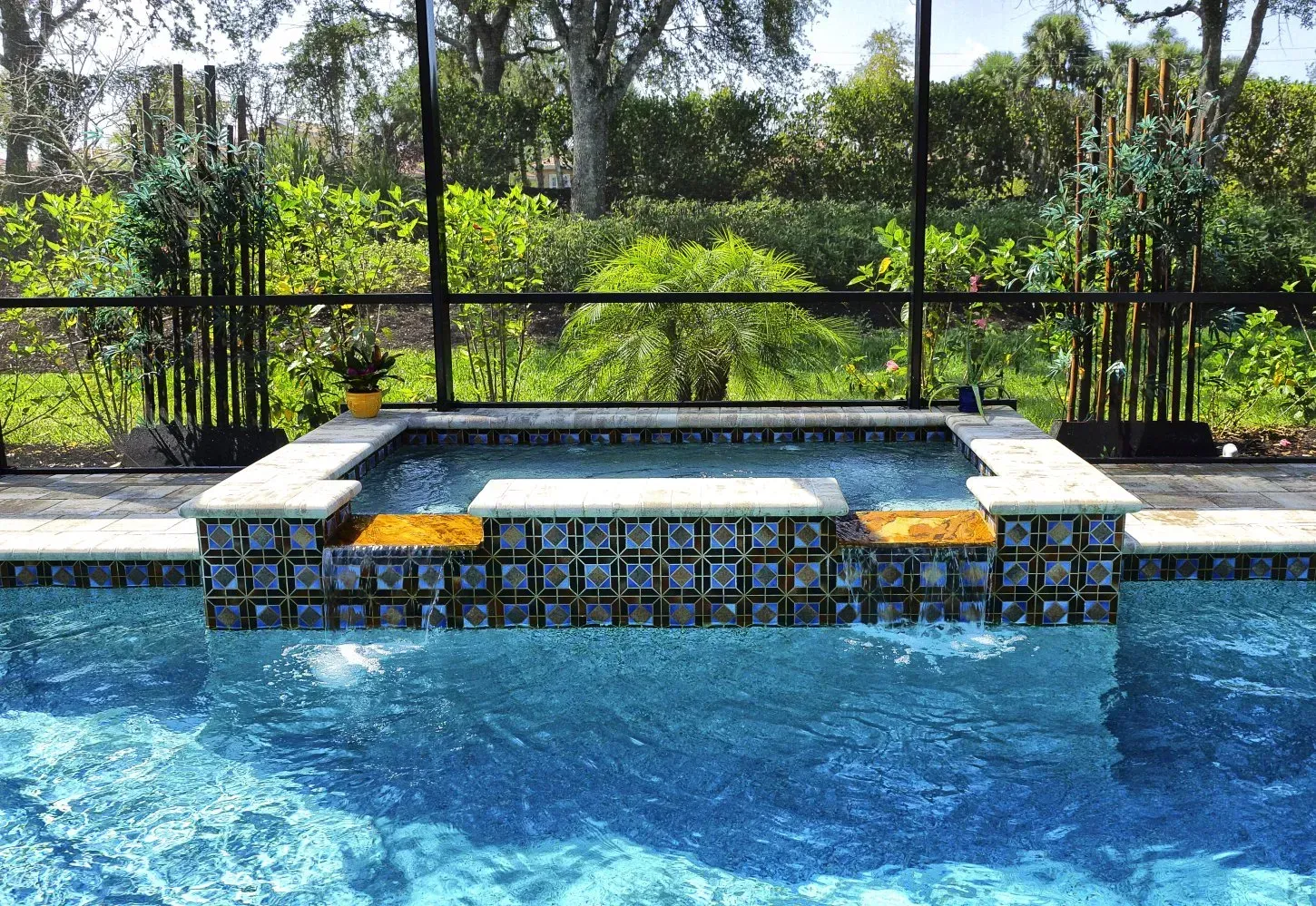 Swimming pool with raised spa, tiled in blue and gold, surrounded by a black screen and greenery.