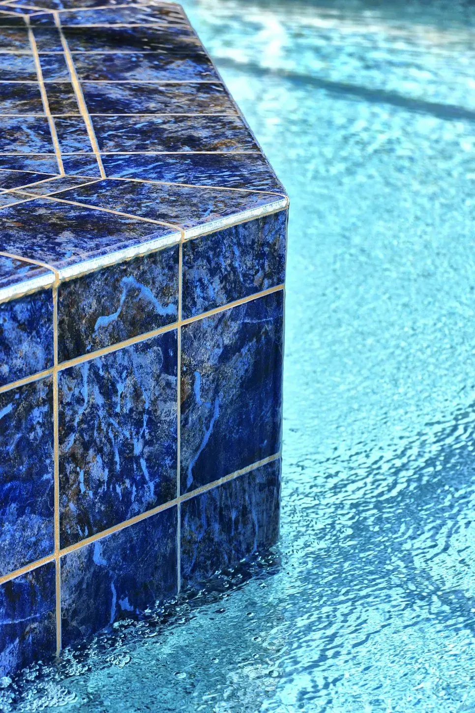 Blue-tiled edge of a pool with shimmering turquoise water, sunlit and vibrant.