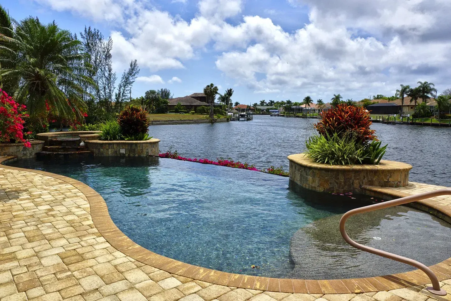 Pool overlooking a canal, with a stone patio, plants, and homes in the background under a cloudy sky.