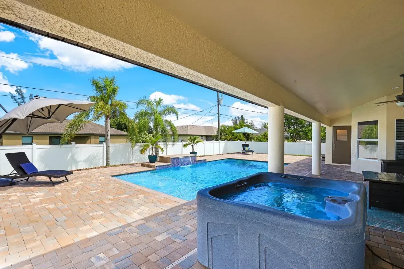 Pool and hot tub under a covered patio; sunny day, blue water, palm trees.