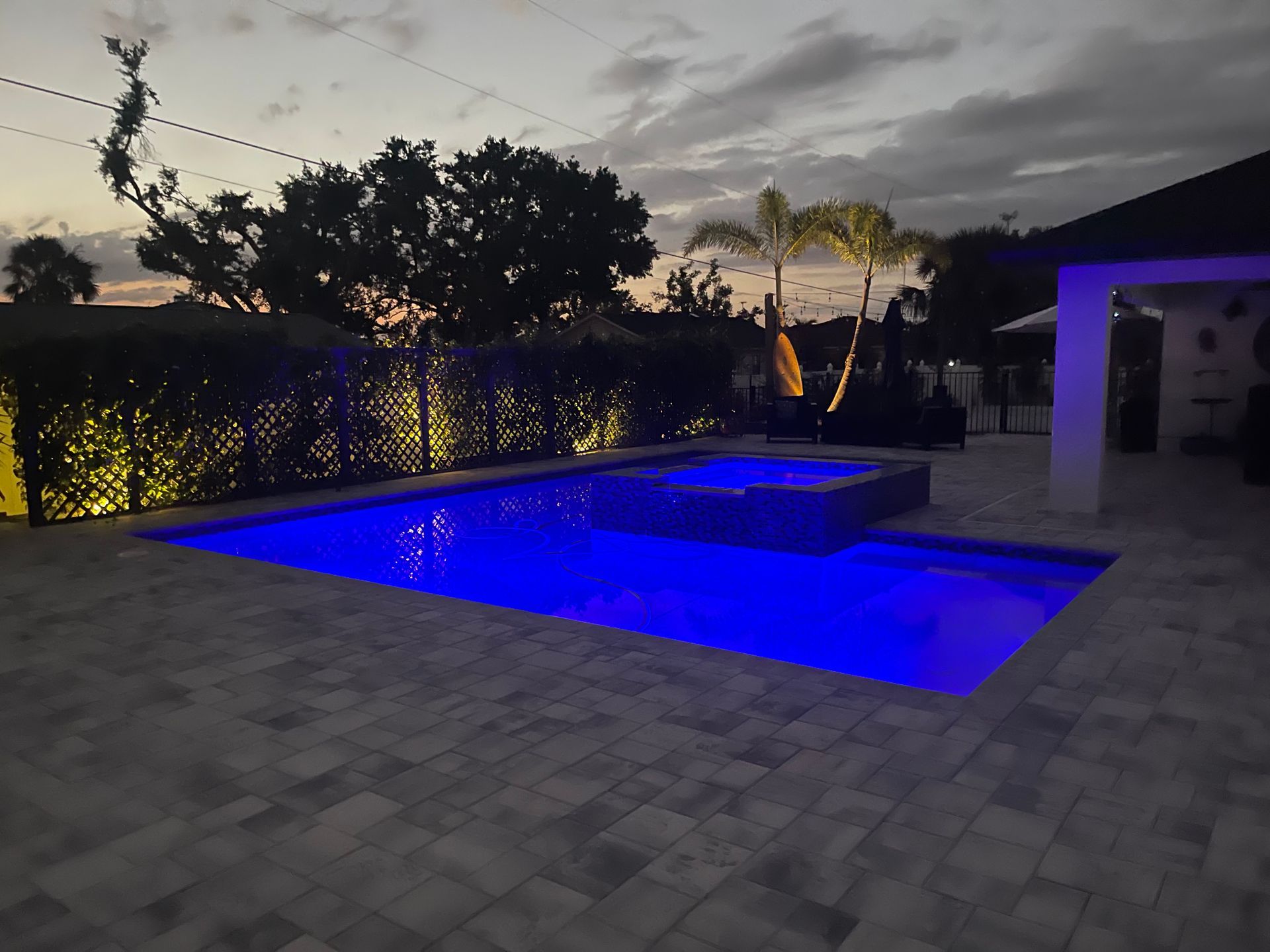 Blue-lit pool and spa at dusk, surrounded by a tiled patio, hedges, and a house.