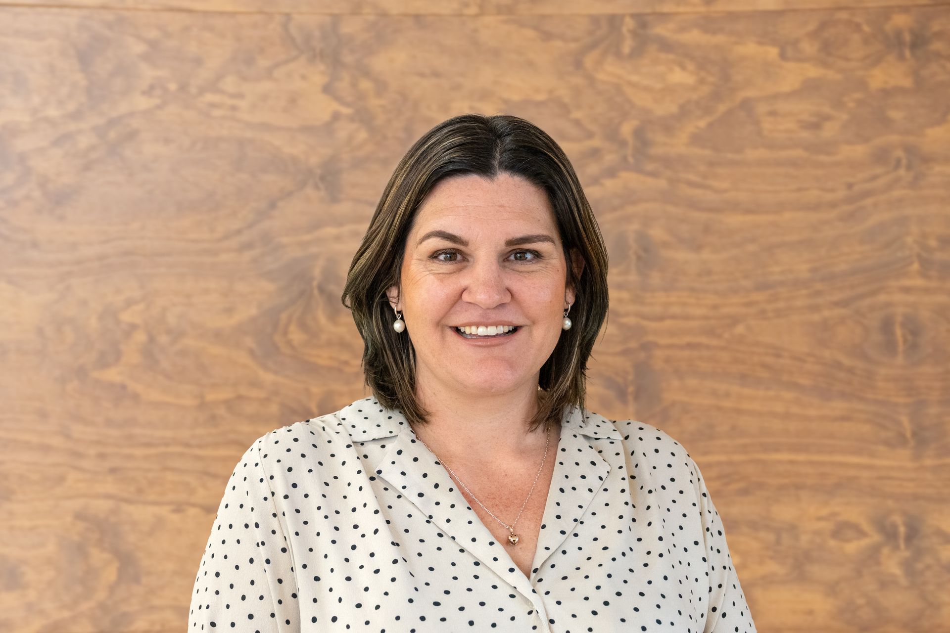 A woman in a polka dot shirt is smiling in front of a wooden wall.