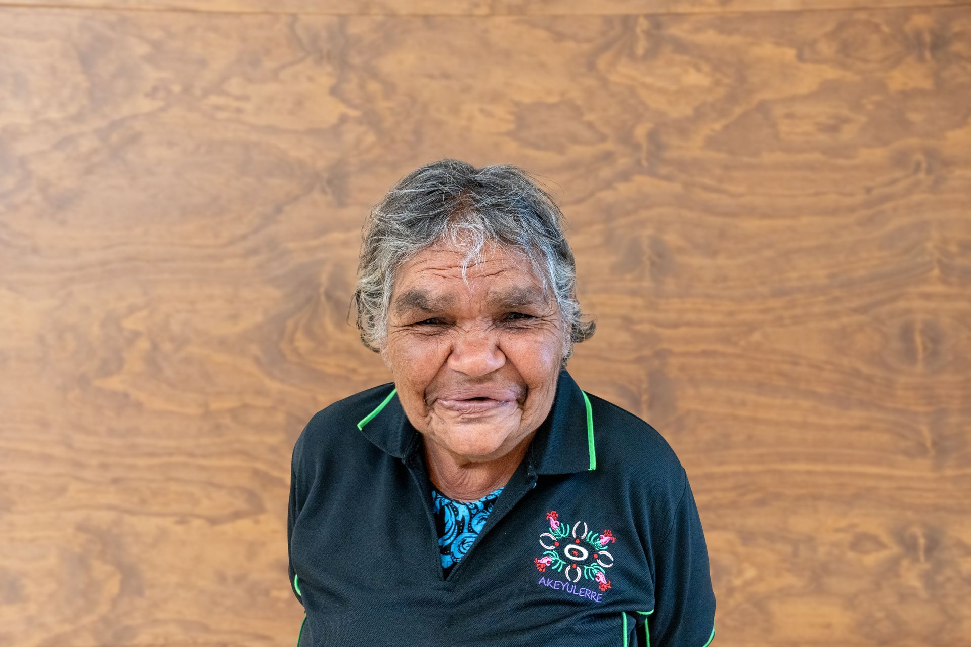 An elderly woman in a black shirt is smiling in front of a wooden wall.