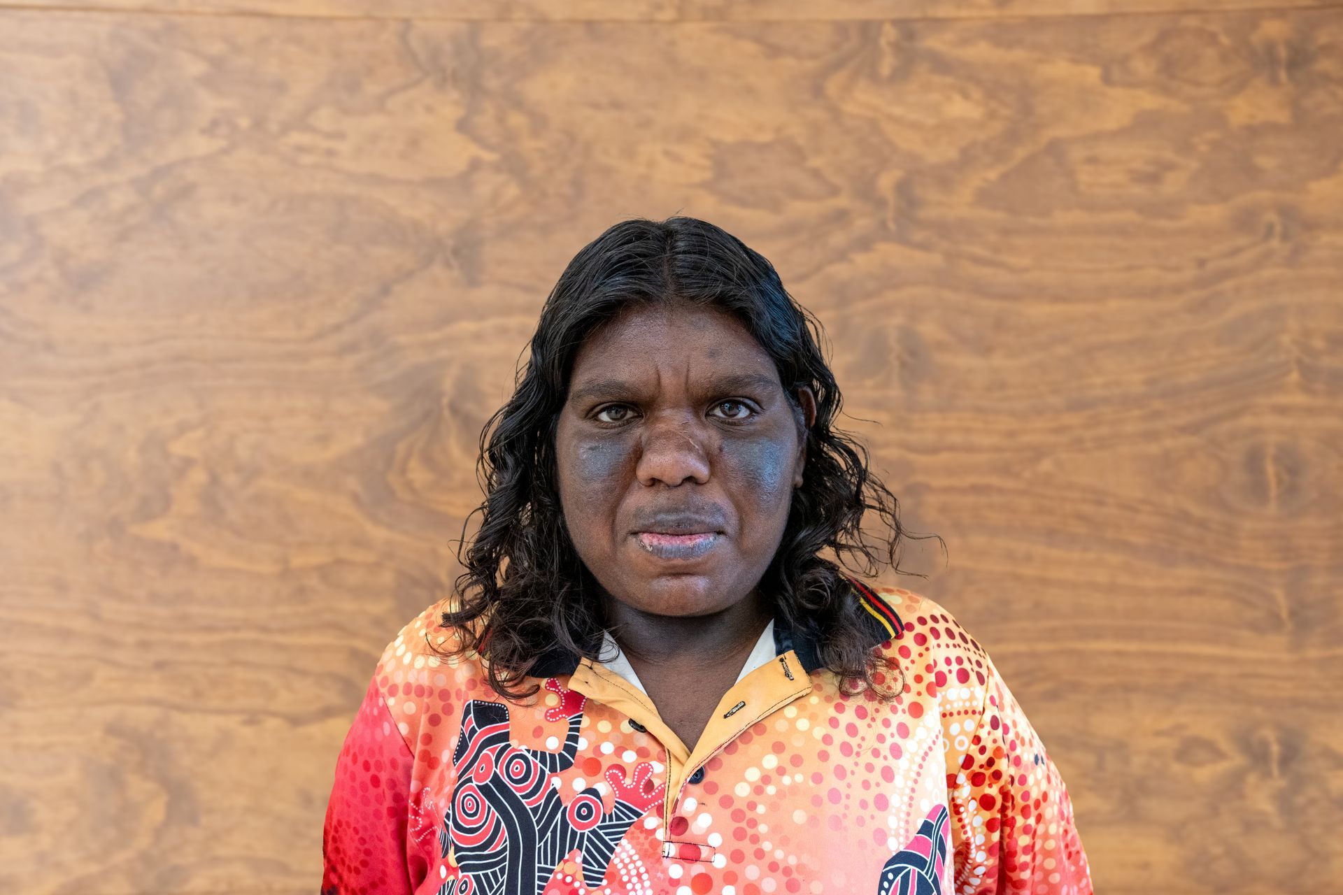 A woman in a colorful shirt is standing in front of a wooden wall.