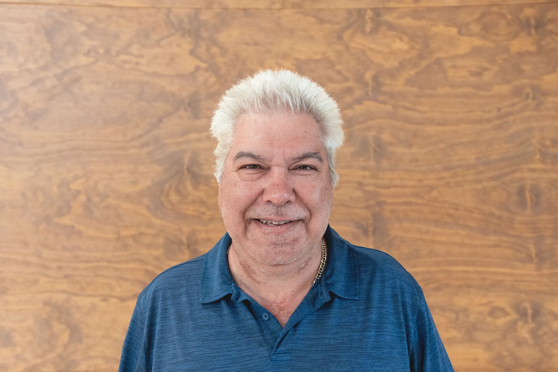 A man in a blue shirt is smiling in front of a wooden wall.