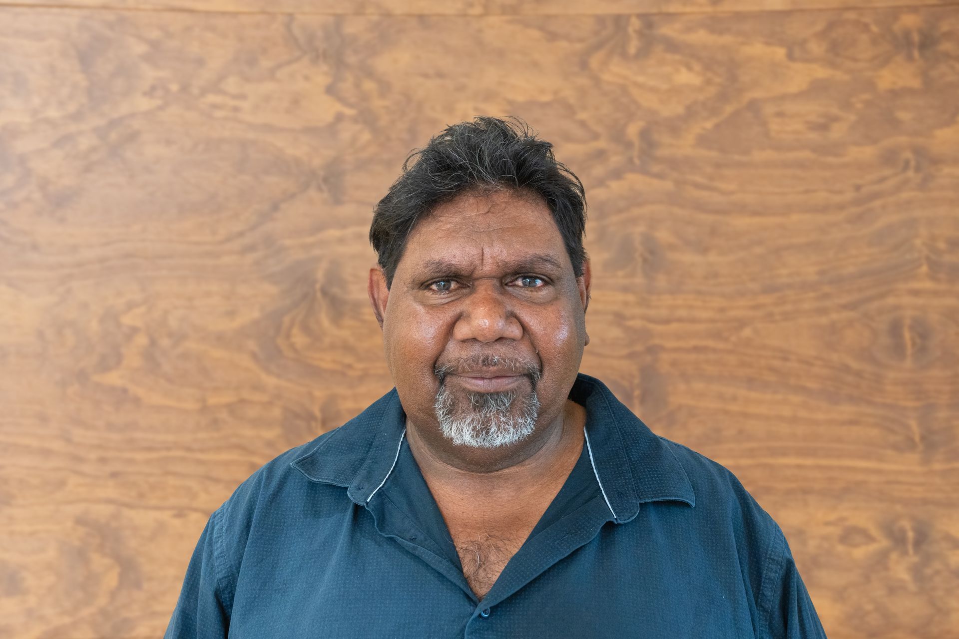 A man with a beard is standing in front of a wooden wall.