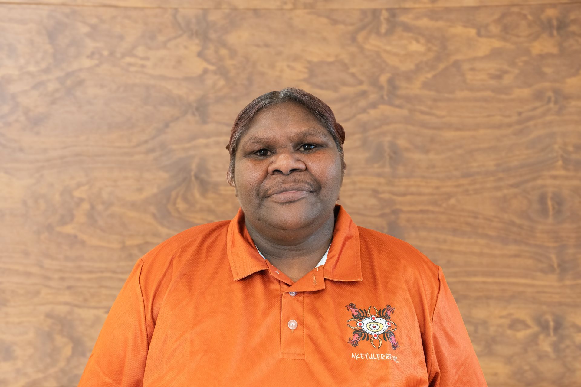 A woman in an orange shirt is standing in front of a wooden wall.