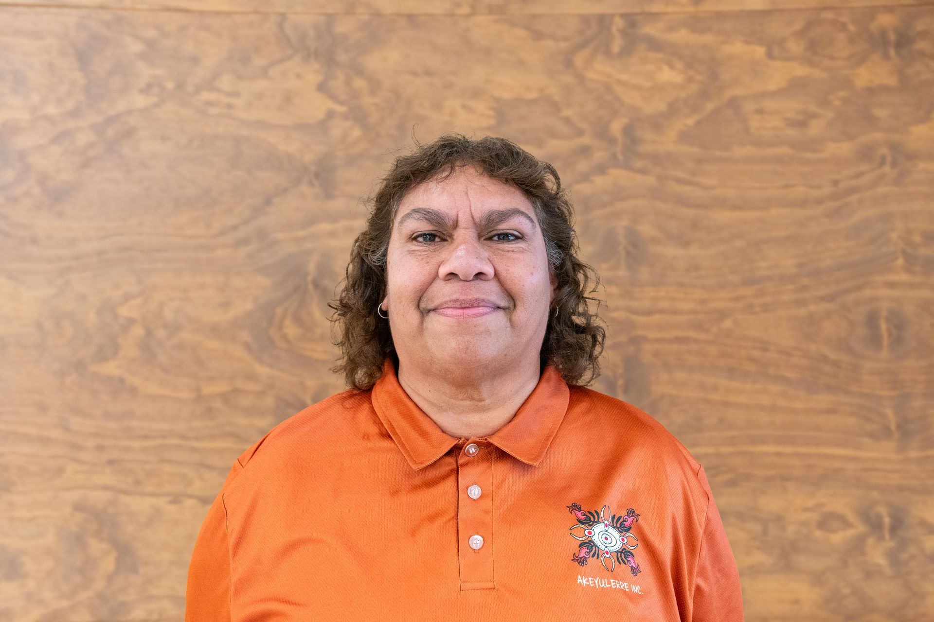 A woman in an orange shirt is smiling in front of a wooden wall.