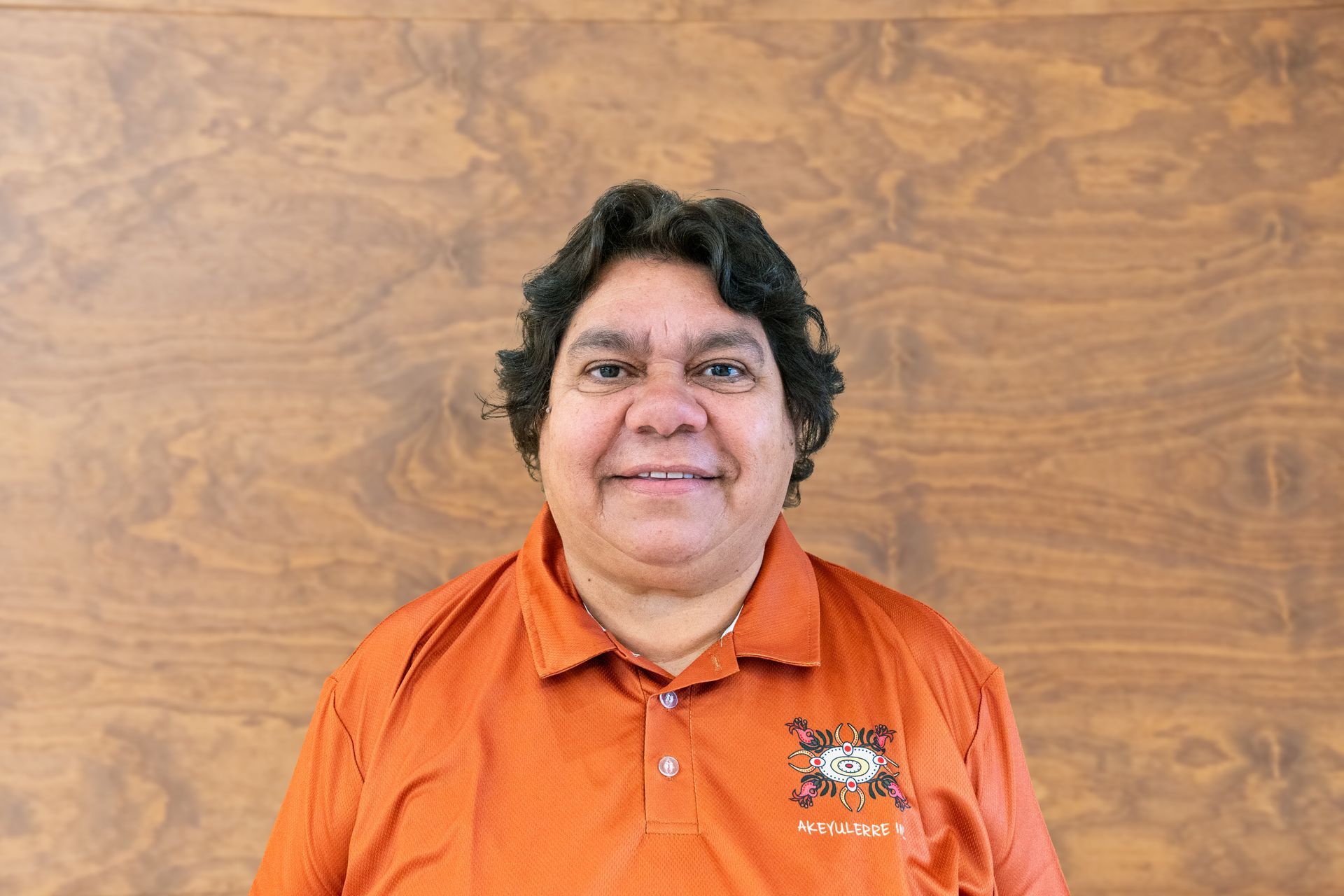 A man in an orange shirt is smiling in front of a wooden wall.