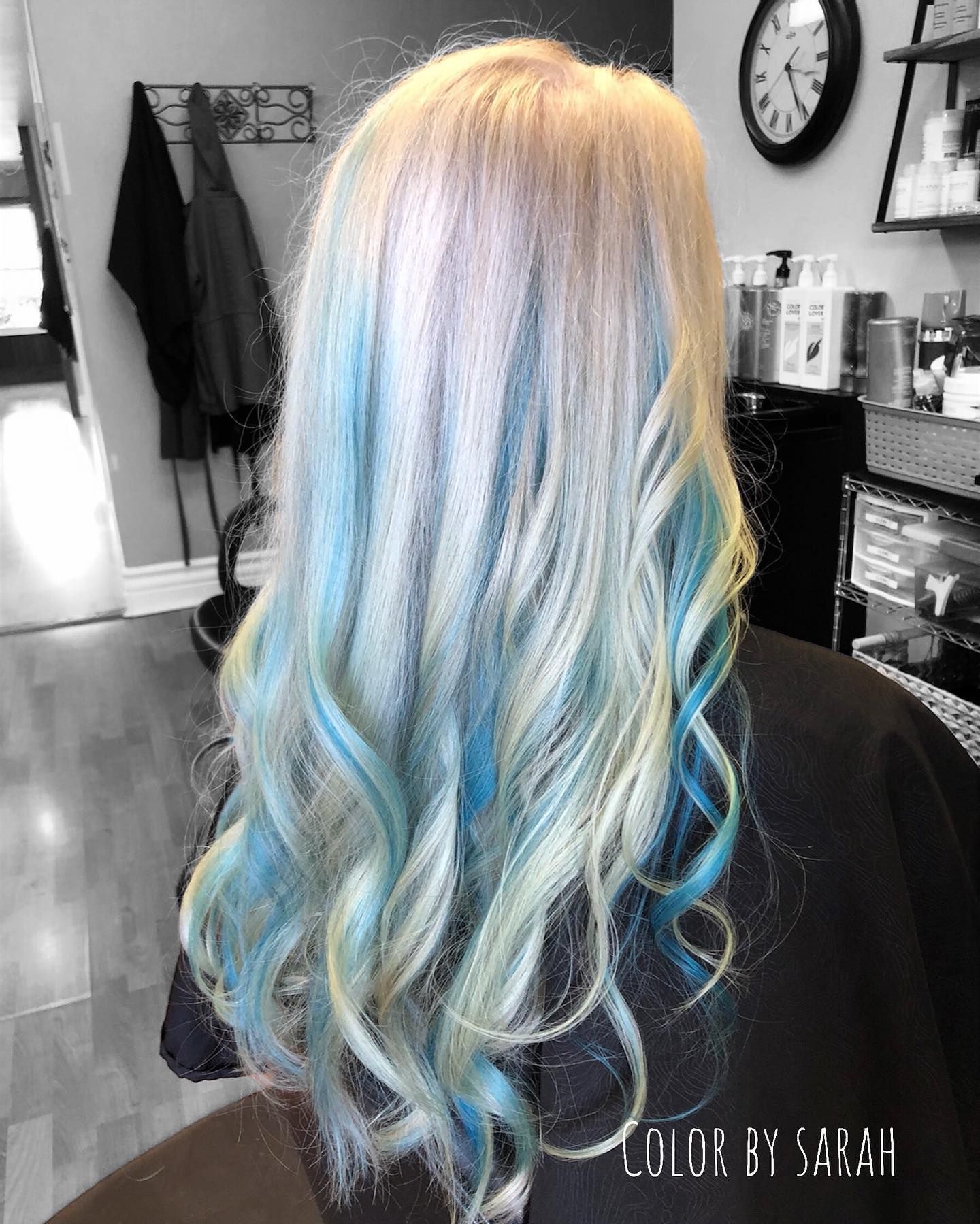 Long wavy blonde hair with pastel blue highlights, styled in a salon chair