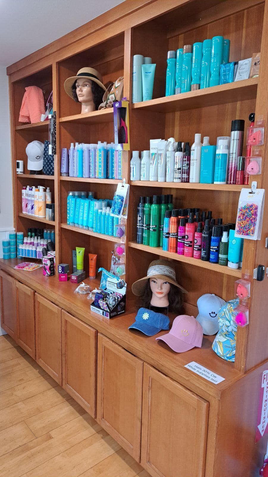 Wooden shelving unit filled with colorful hair products and salon displays, with mannequin heads and hats.