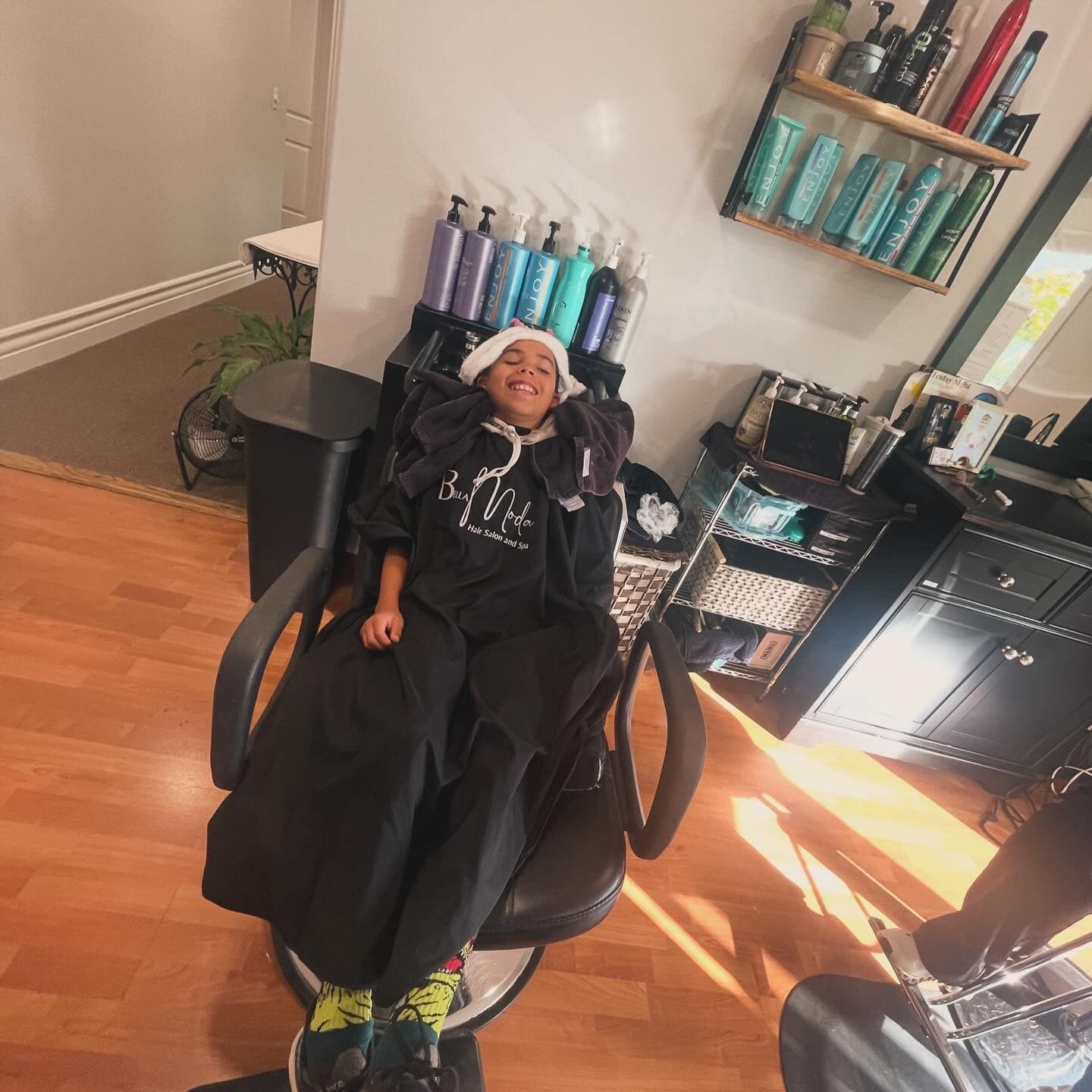 Person reclining in a salon chair, wearing black, with colorful hair products and tools on shelves nearby.