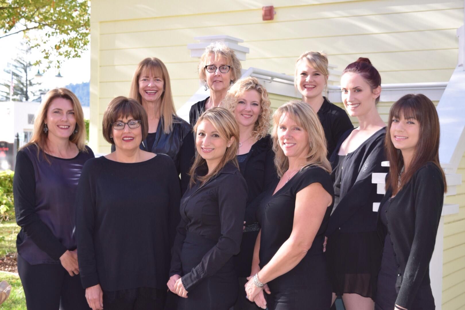 Nine women in black outfits posing together outdoors in front of a light-colored building
