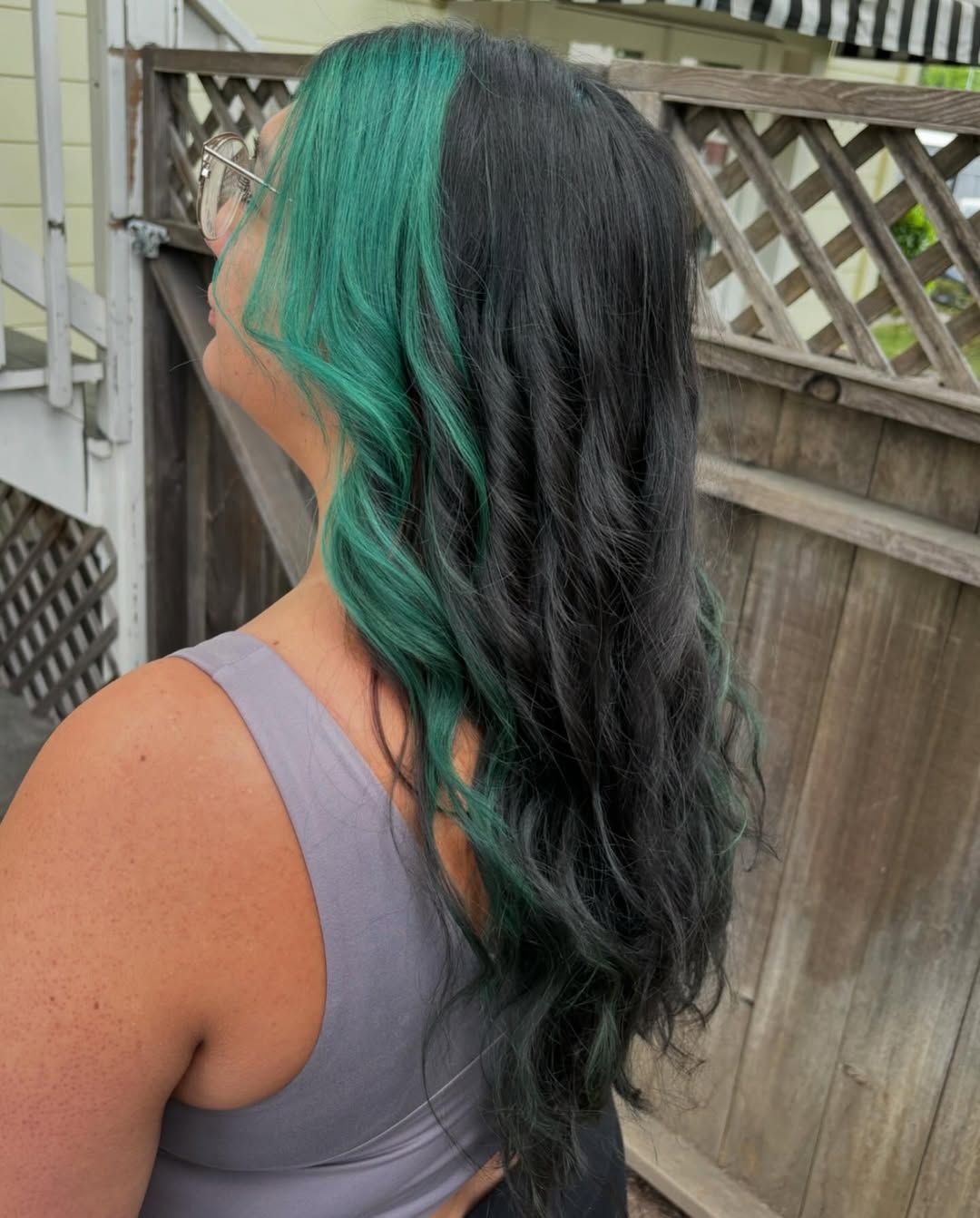 Side view of long black hair with green highlights, worn with a gray tank top outdoors by a wooden fence
