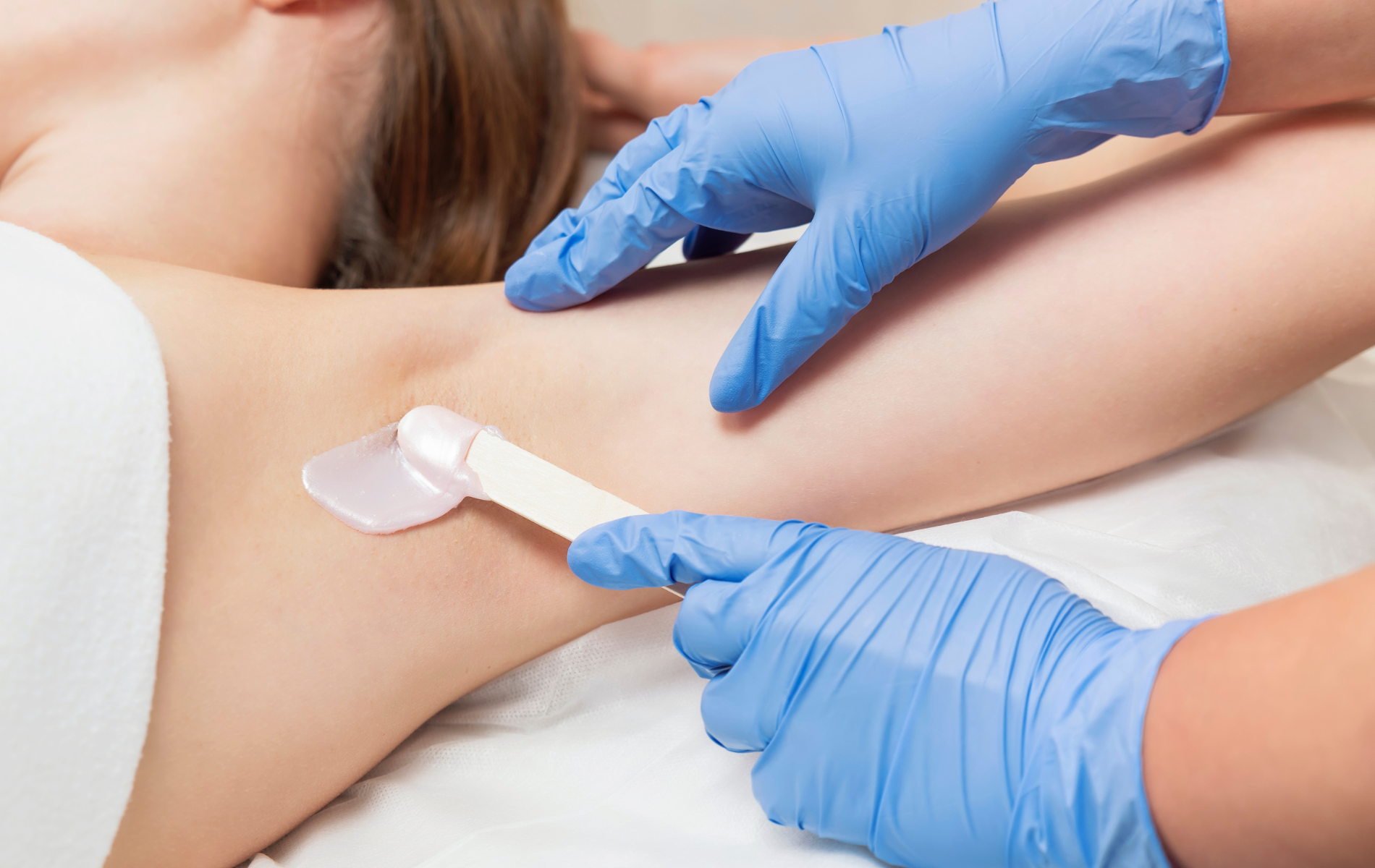 Gloved hands waxing a person’s underarm with a spatula in a spa setting