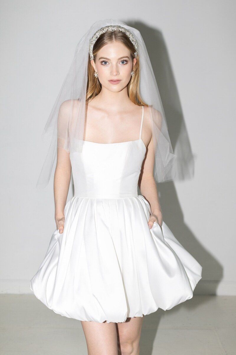 A woman in a white dress and veil is standing in front of a white wall.