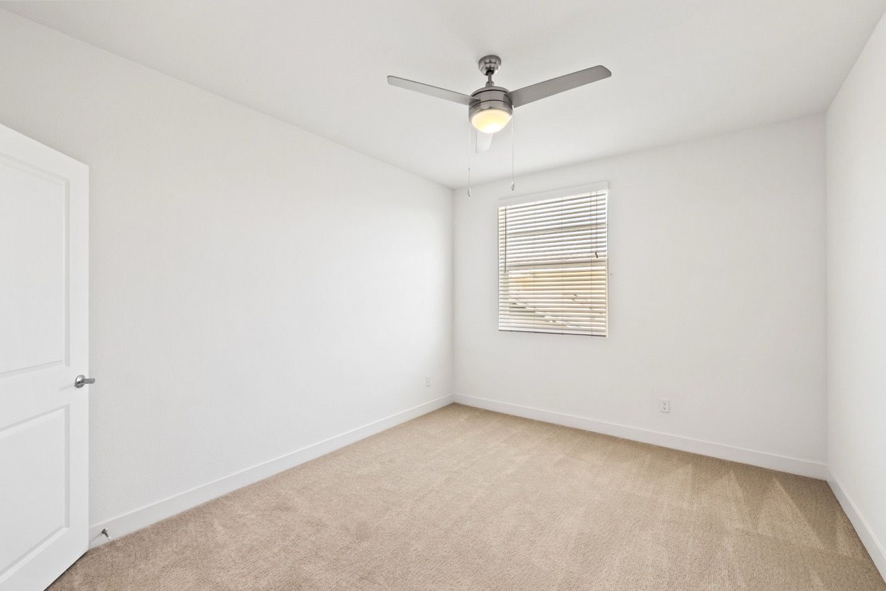 Empty bedroom with beige carpet, white walls, a ceiling fan, and a window with blinds.