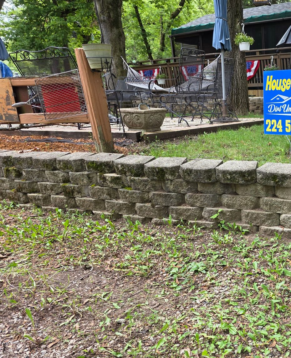 Stone retaining wall in front of a yard with trees, chairs, and a building in the background.