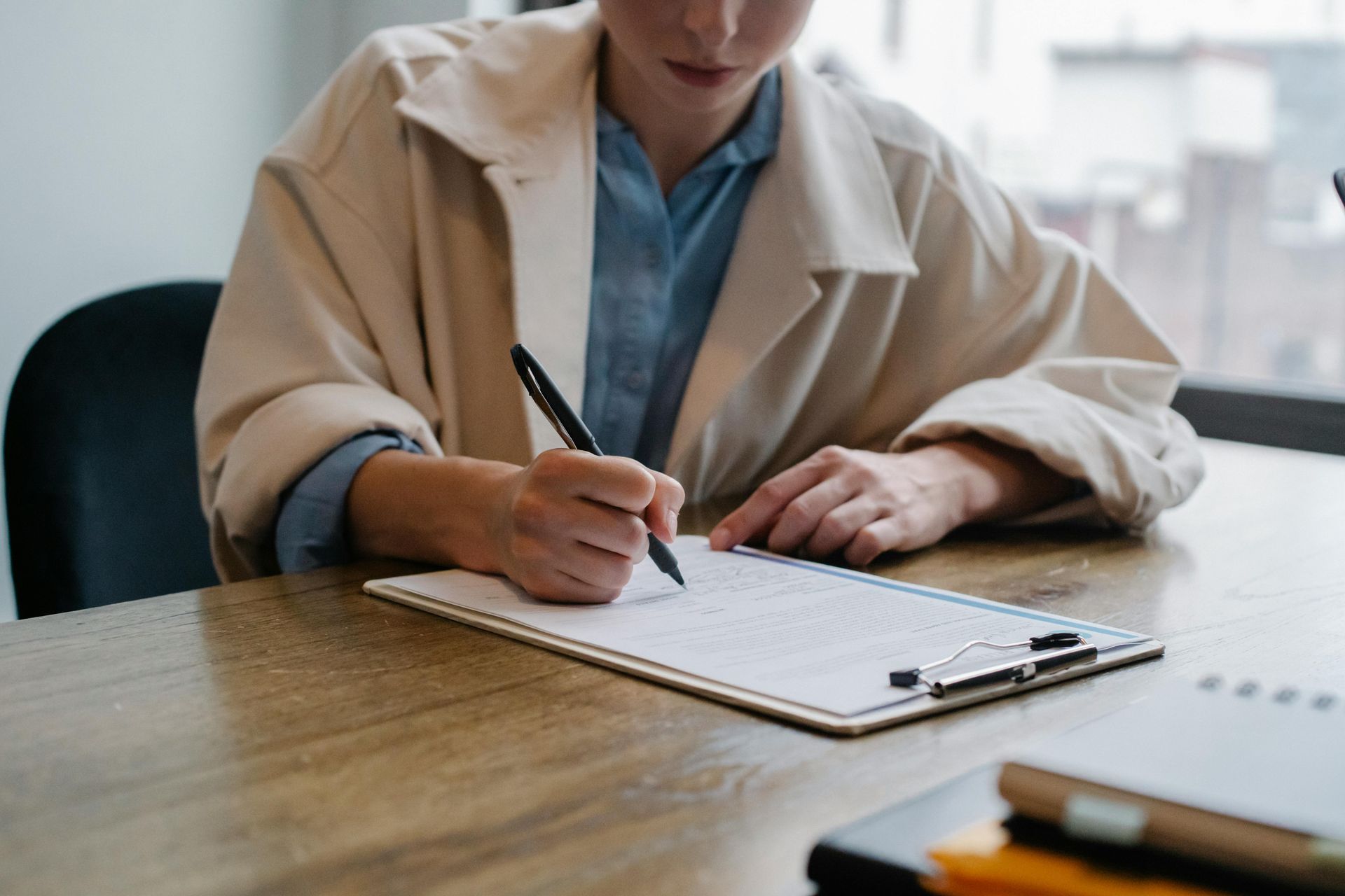 Person writing in a notebook at a wooden desk, wearing a beige coat in a bright office setting