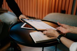 Two people using a tablet at a small round table with a notebook and pen nearby