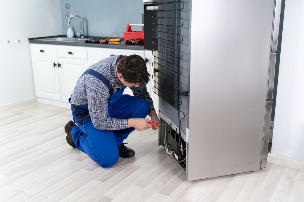 A man is kneeling down to fix a refrigerator in a kitchen.
