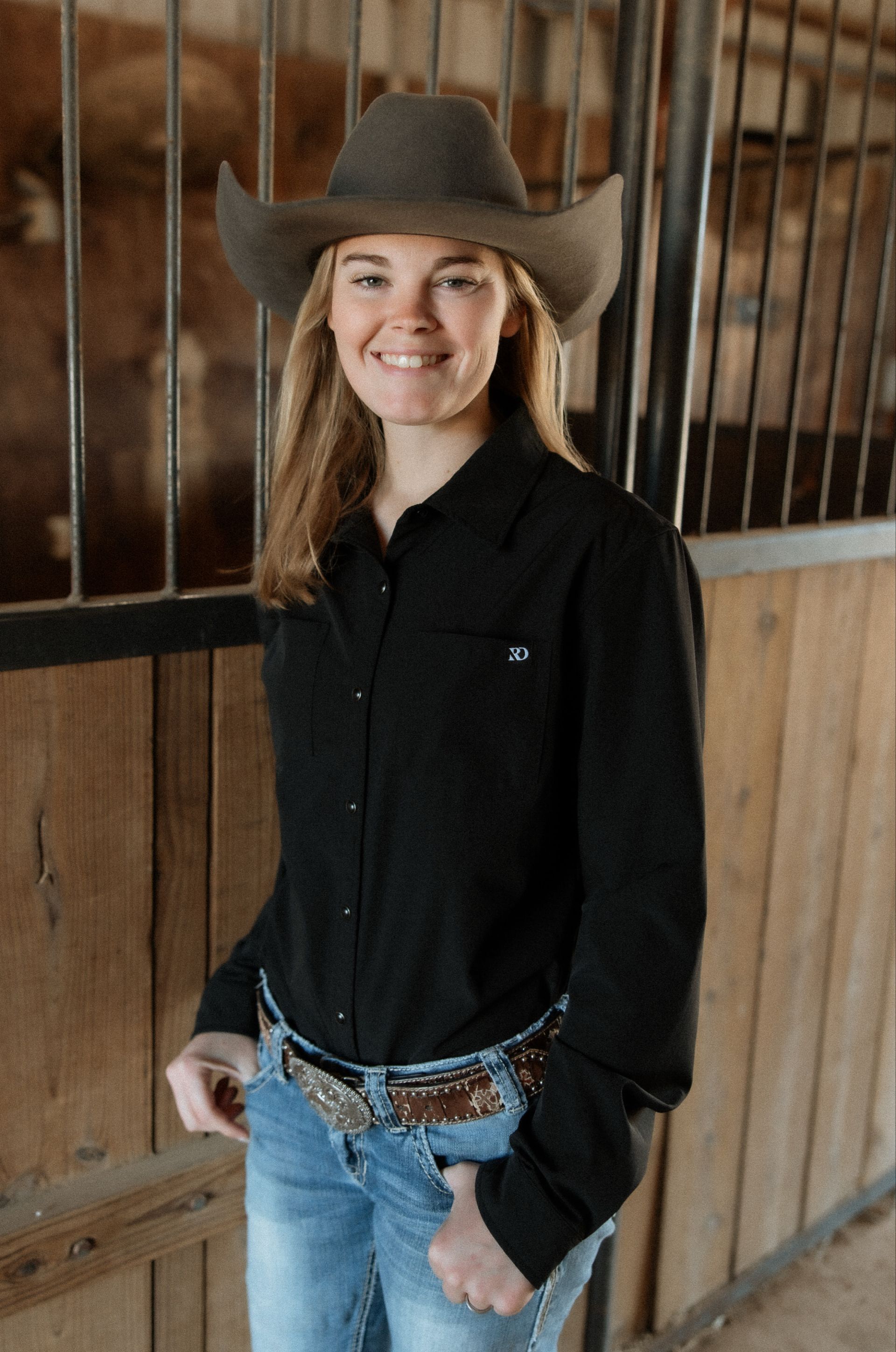 A woman wearing a cowboy hat and a black shirt is standing in a barn.