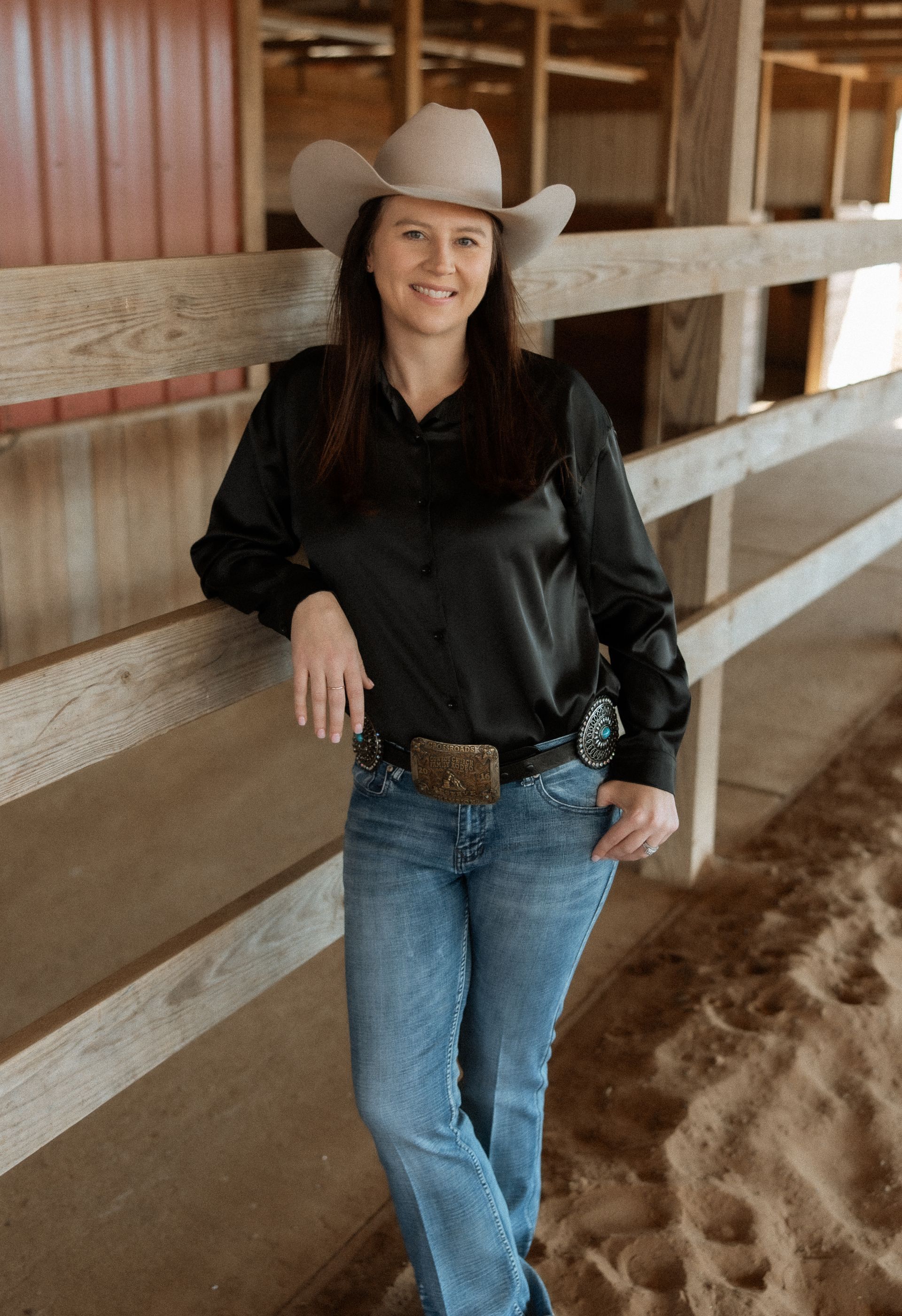 A woman wearing a cowboy hat and jeans is leaning against a fence.