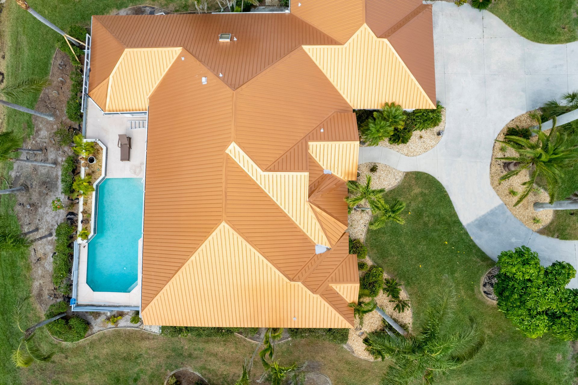 Overhead view of a house with a brown roof, pool, and circular driveway, surrounded by green grass and palm trees.
