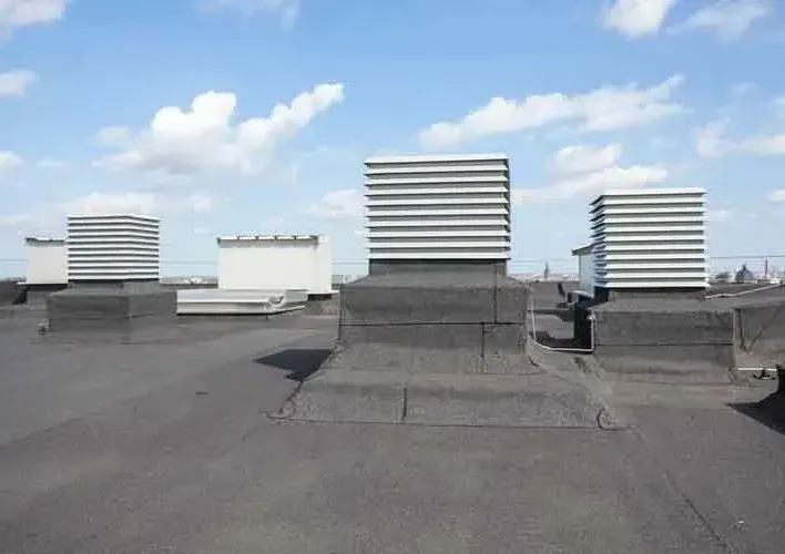 Flat rooftop with several square, louvered ventilation units on a black tar surface, under a blue sky with clouds.